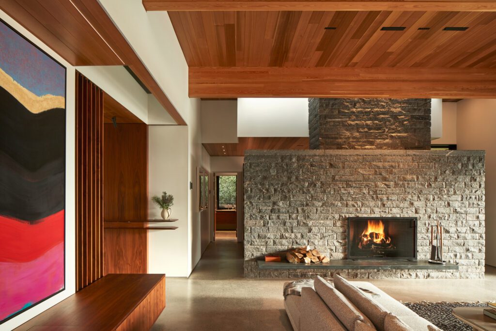 Great room of a modern home with exposed wooden beams, walnut casework, and stone fireplace