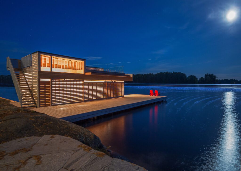 Vista exterior nocturna de un moderno cobertizo para botes prefabricado de madera, iluminado desde dentro, en un lago