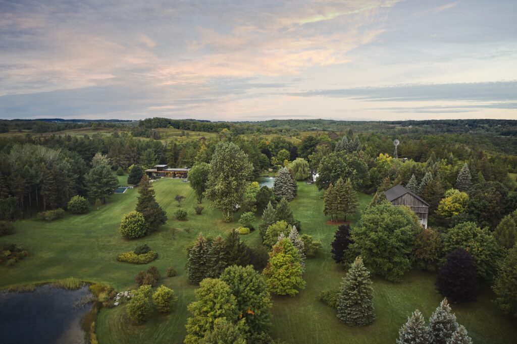 Aerial view of a wooded landscape with trees and a small body of water. A modern house is visible near a historic wooden building.