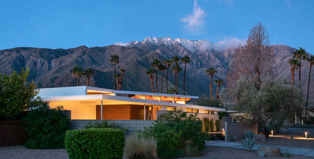 View of a modern post-and-beam prefab home at twilight backed by palm trees and snow-capped mountains