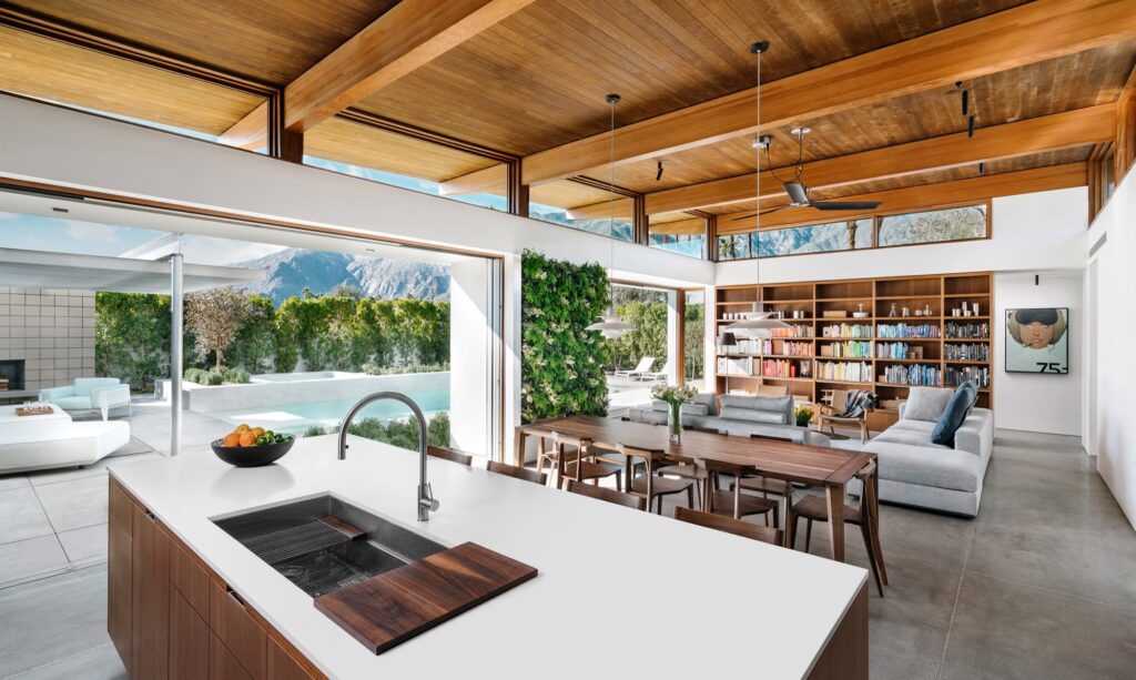 Interior shot of the great room of a modern prefab custom home, open to the courtyard and pool, with a kitchen island, dining table, couches, and bookshelves. Strong wooden beams traverse the ceiling.