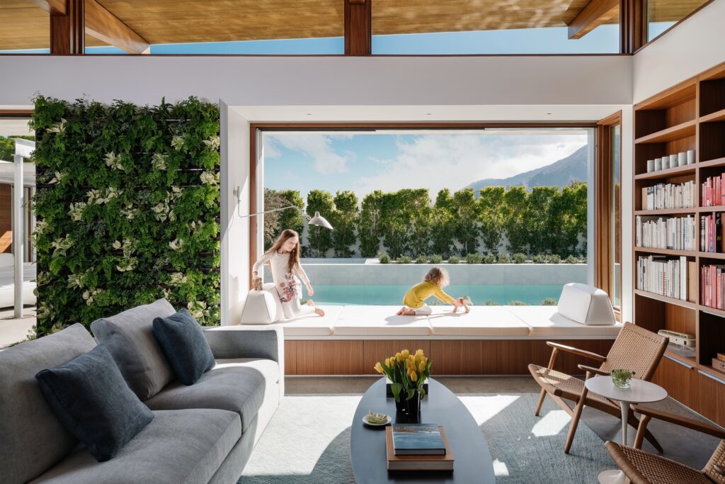View from the great room of a modern custom prefab home out to the pool, with two girls playing on the living platform by the window