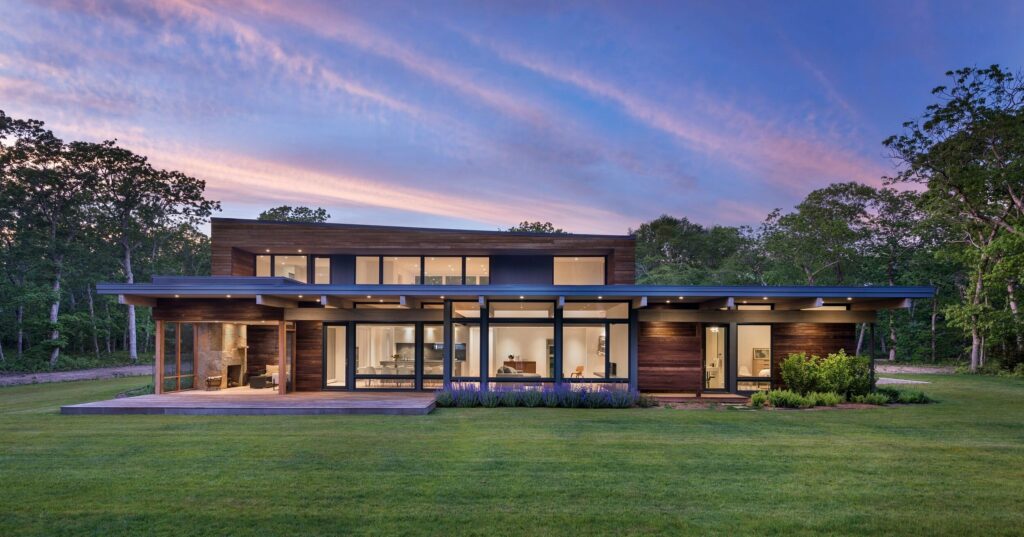 Modern home at dawn with a double-height great room, lit from within, on a green lawn