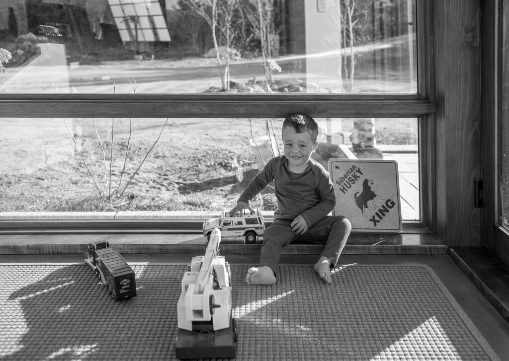 Black and white image of a boy playing with a truck in front of the large window of a home