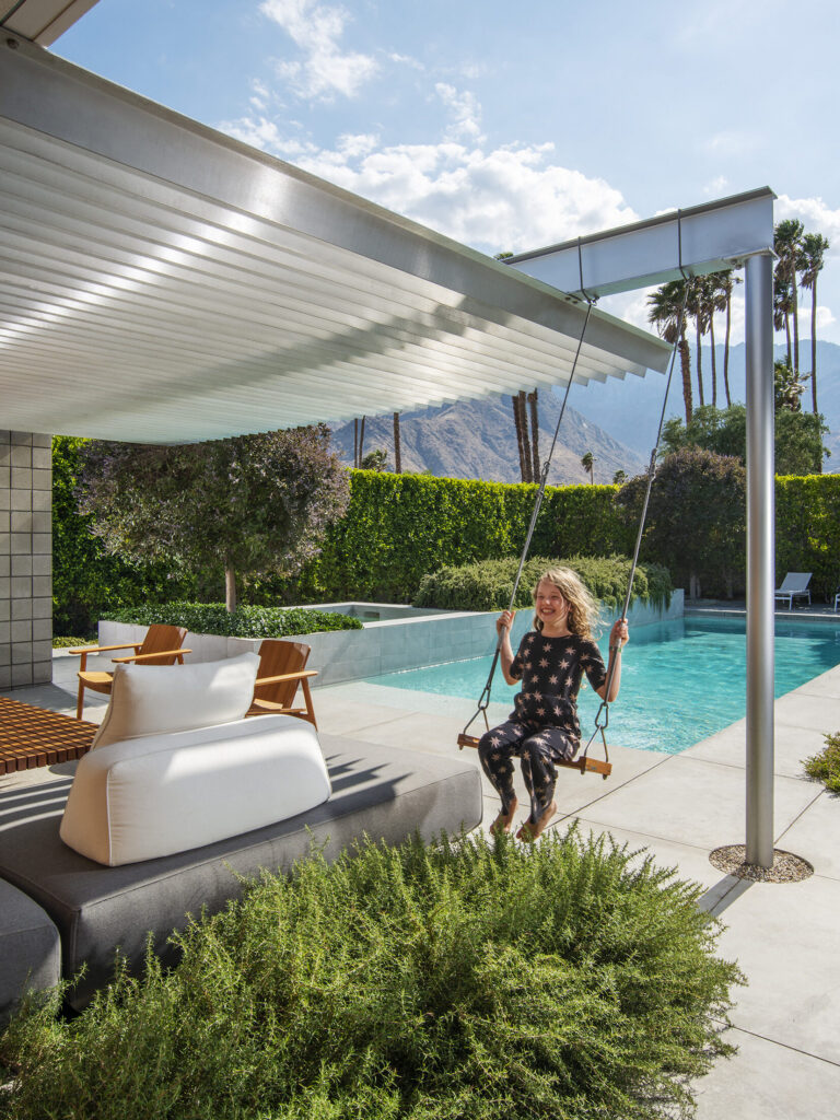 A girl on a swing under a metal pergola on a patio with a pool, a mountain, and palm trees in the background