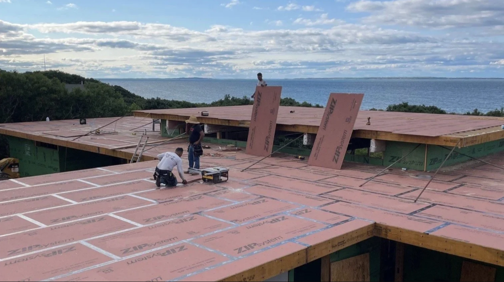 A small crew works on the under-construction roof of a modern home overlooking the ocean